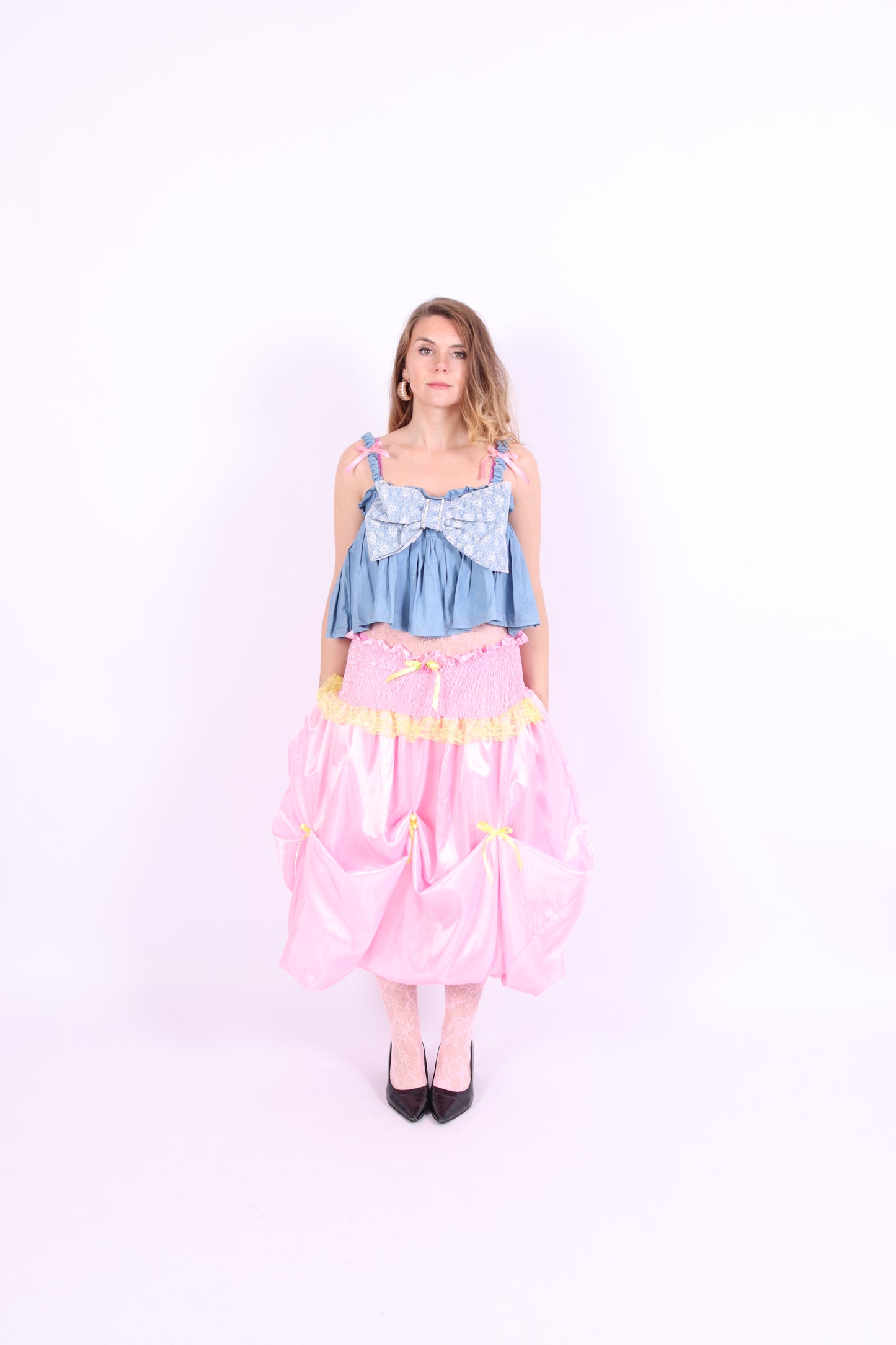 A denim crop top with a large white lace bow and diamante detail on the chest, pink topstitch elasticated shirring across the top bodice, and full babydoll gathered panel. It features elasticated shoulder straps with pink ribbon bow detail. Delta Of Phoenix Gigi Bow Top