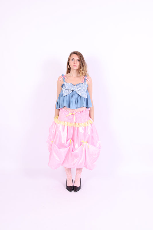 A denim crop top with a large white lace bow and diamante detail on the chest, pink topstitch elasticated shirring across the top bodice, and full babydoll gathered panel. It features elasticated shoulder straps with pink ribbon bow detail. Delta Of Phoenix Gigi Bow Top