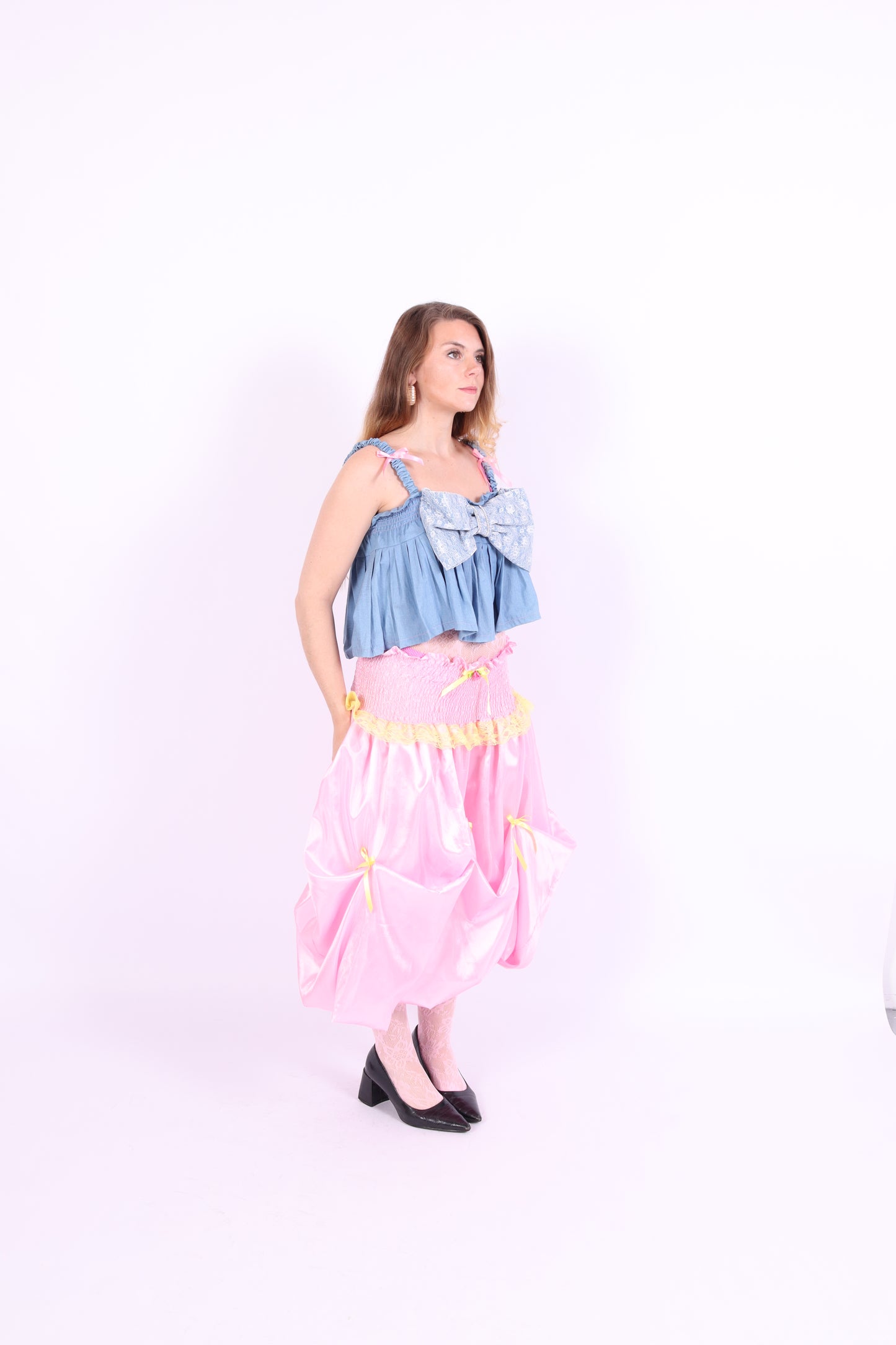 A denim crop top with a large white lace bow and diamante detail on the chest, pink topstitch elasticated shirring across the top bodice, and full babydoll gathered panel. It features elasticated shoulder straps with pink ribbon bow detail. Delta Of Phoenix Gigi Bow Top