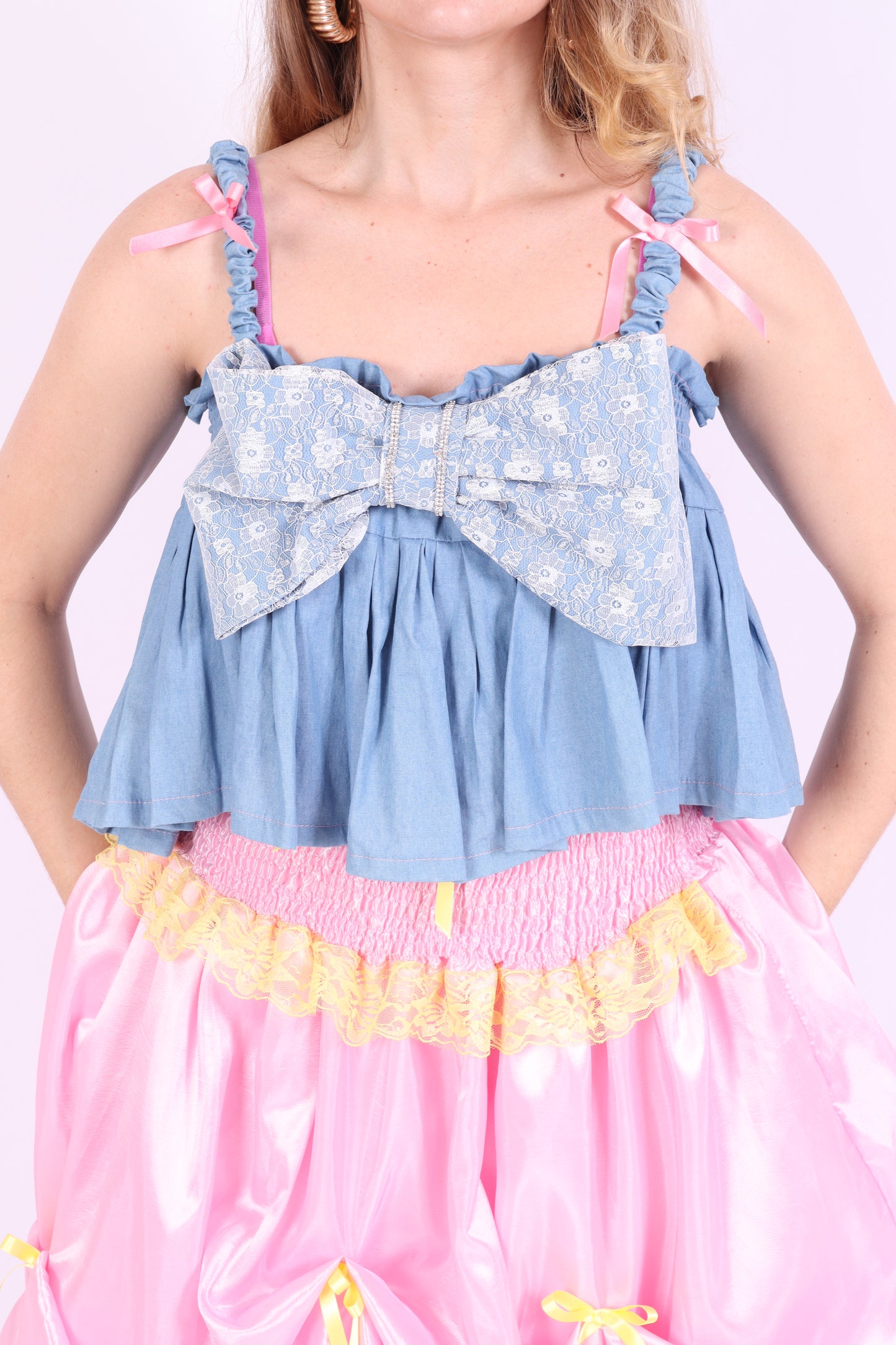 A denim crop top with a large white lace bow and diamante detail on the chest, pink topstitch elasticated shirring across the top bodice, and full babydoll gathered panel. It features elasticated shoulder straps with pink ribbon bow detail. Delta Of Phoenix Gigi Bow Top