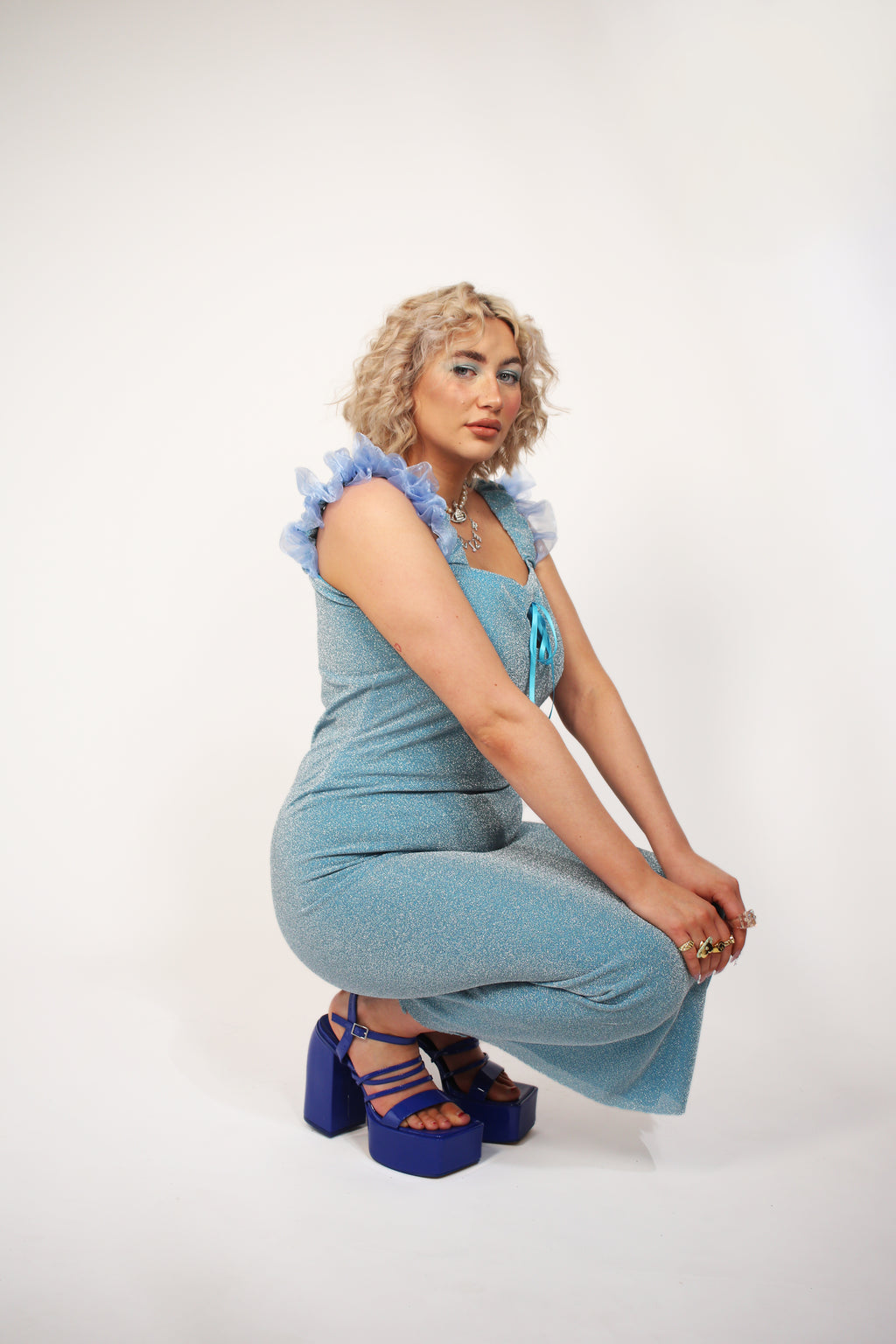 A blue glitter cocktail dress with ruffle detailing, elasticated ruched straps, and a ribbon bow keyhole detail at the front. Delta Of Phoenix Juni Midi Dress