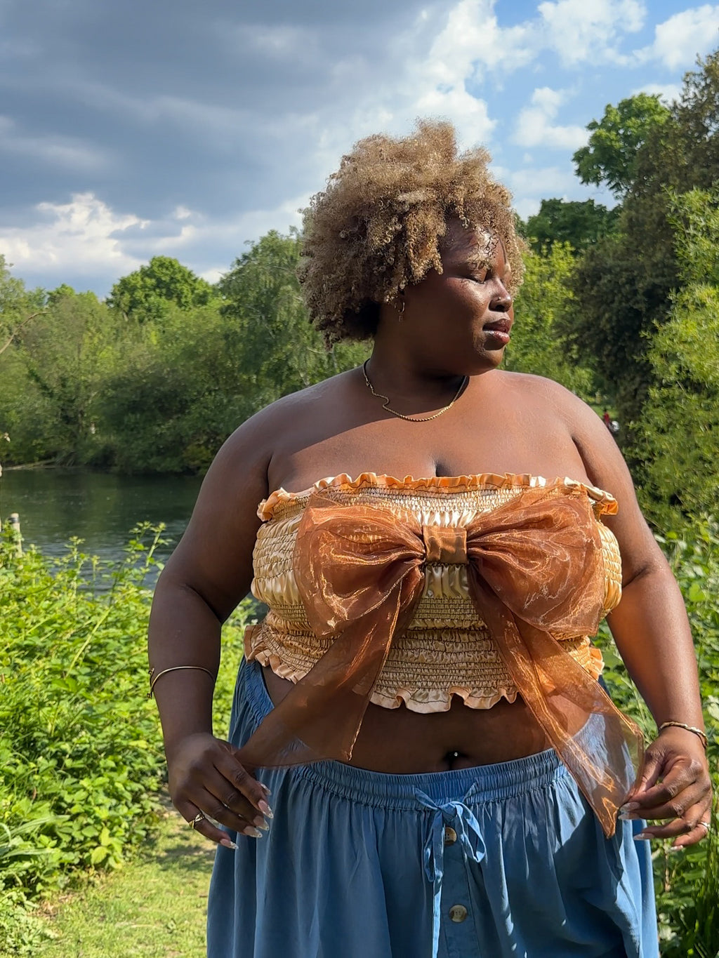 A gold satin top with an elasticated shirring bodice and a terracotta brown organza bow on the front. It features shoulder straps with elasticated and ruched details. Delta Of Phoenix Naomi Bow Top - worn by Naomi Native