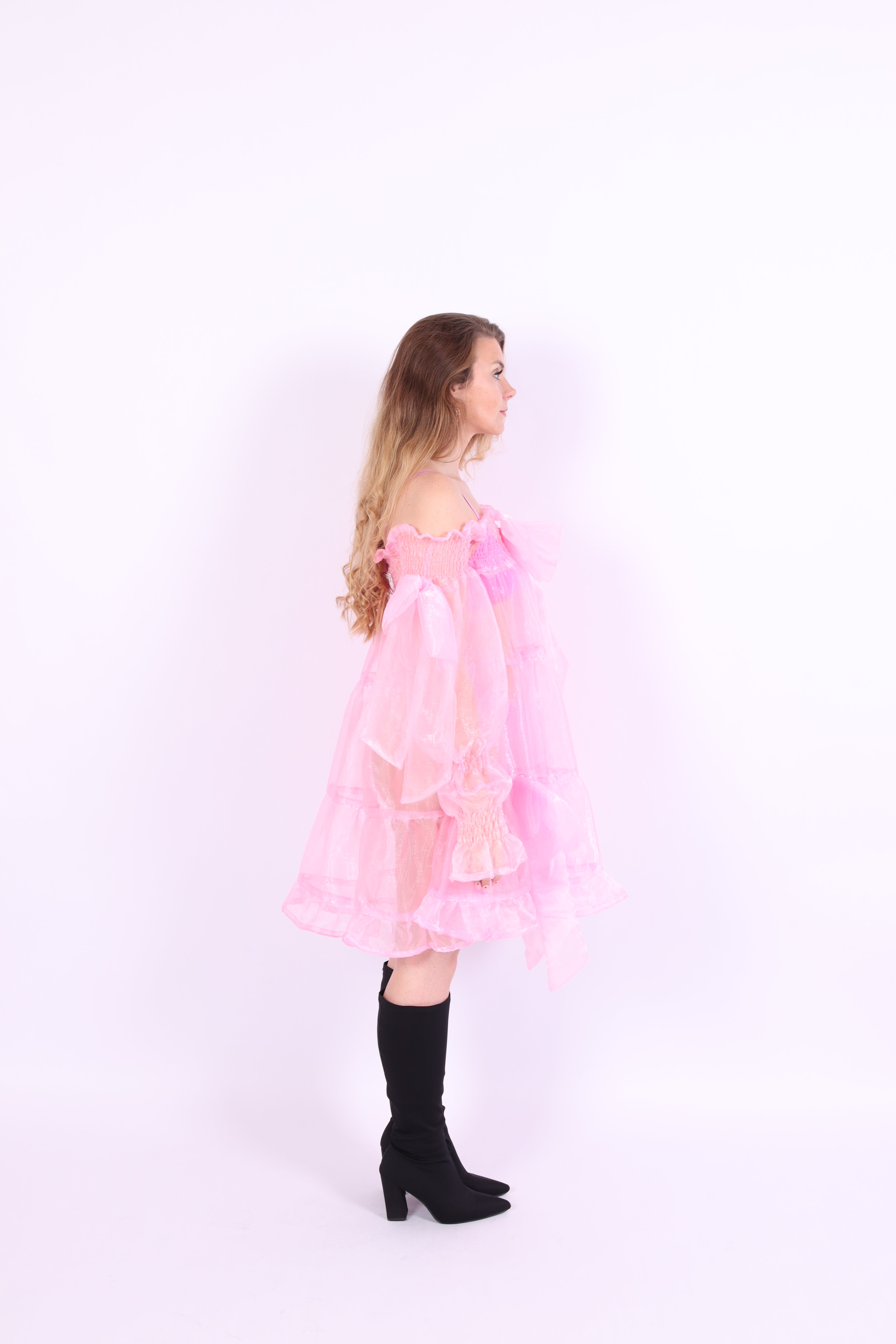A bubblegum pink organza mini dress with a large bow detail at the sleeve, skirt, and front, featuring a bardot neckline and long puff sleeves. Cosette Dress by Delta Of Phoenix