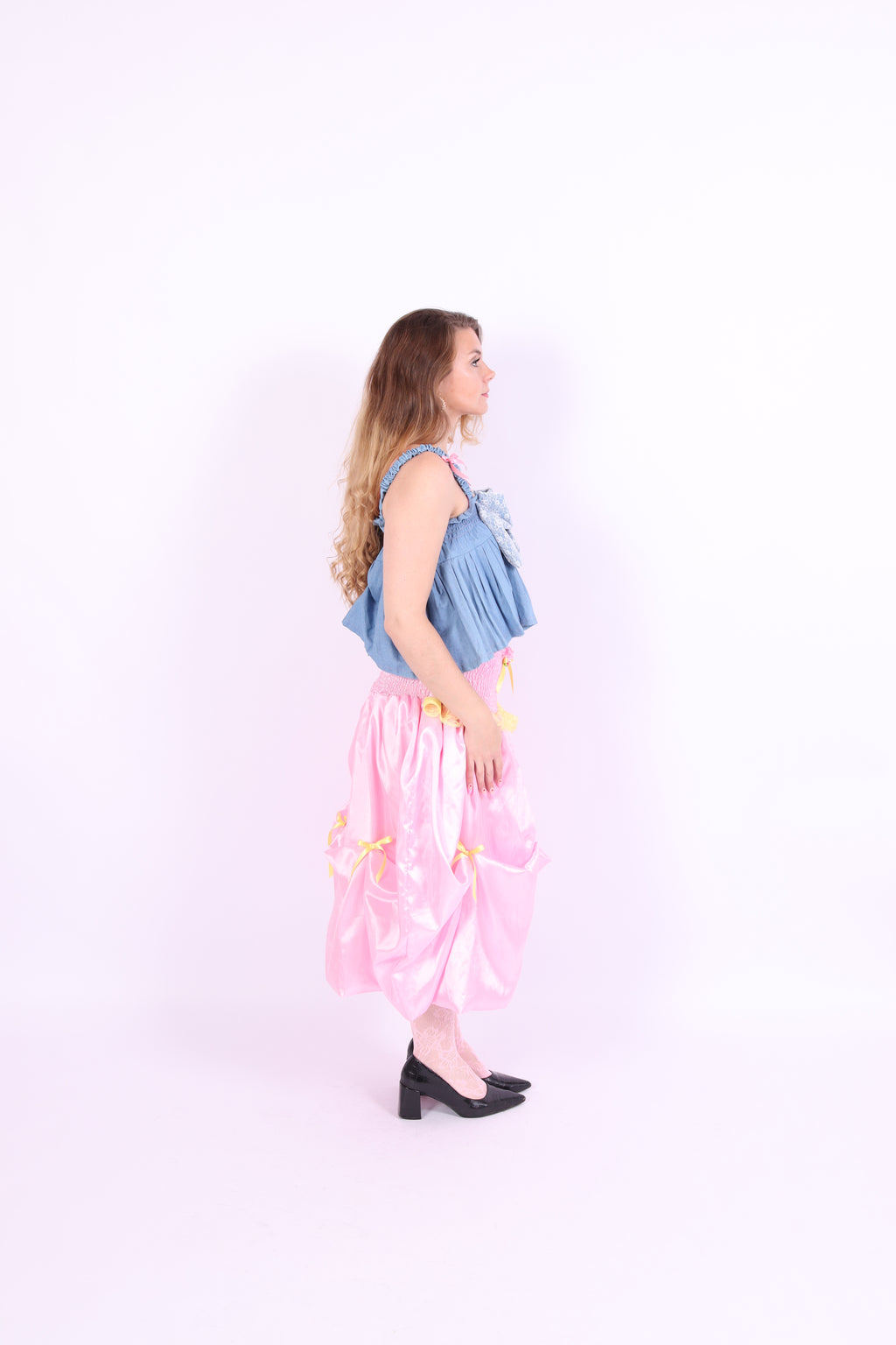 A denim crop top with a large white lace bow and diamante detail on the chest, pink topstitch elasticated shirring across the top bodice, and full babydoll gathered panel. It features elasticated shoulder straps with pink ribbon bow detail. Delta Of Phoenix Gigi Bow Top