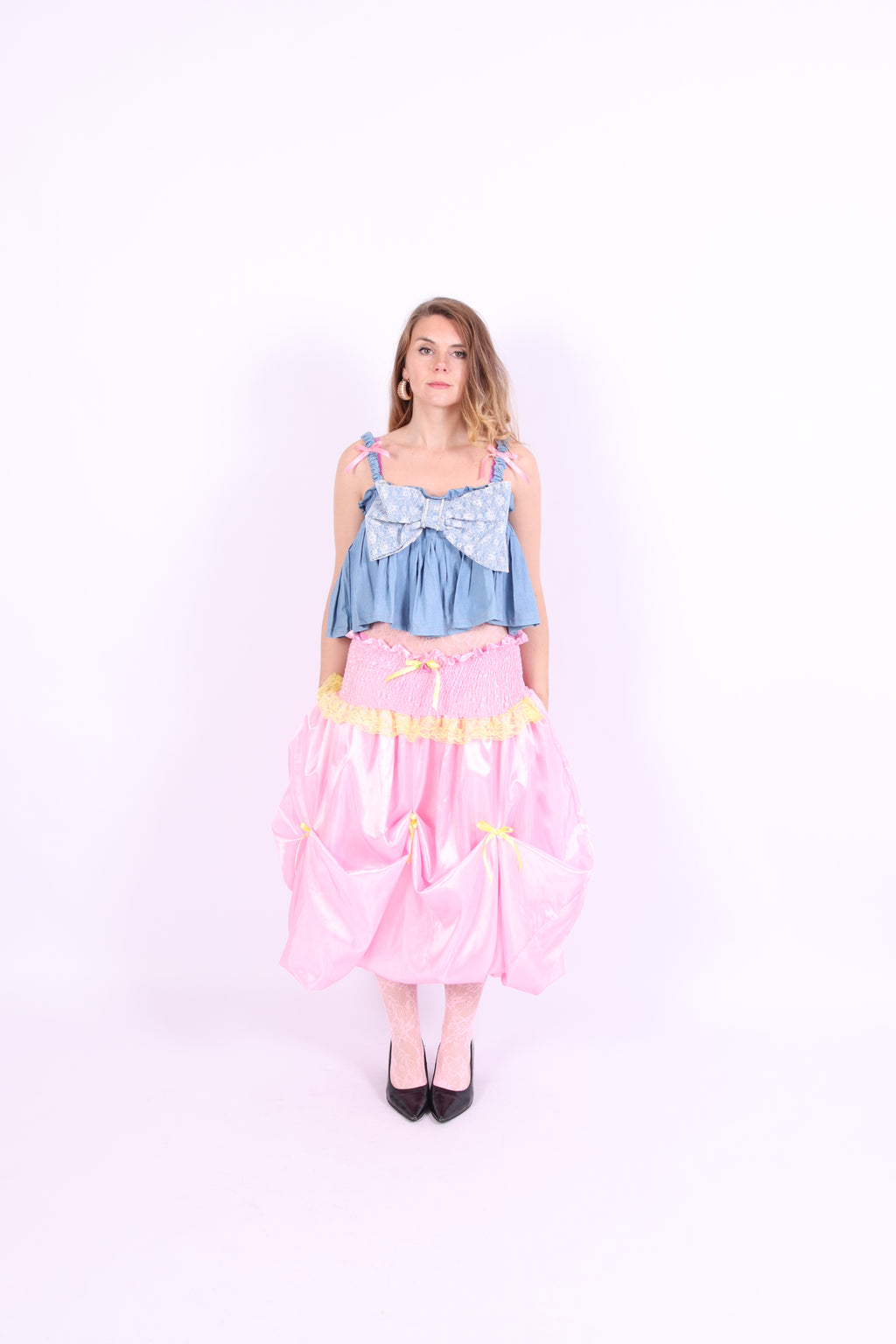 A denim crop top with a large white lace bow and diamante detail on the chest, pink topstitch elasticated shirring across the top bodice, and full babydoll gathered panel. It features elasticated shoulder straps with pink ribbon bow detail. Delta Of Phoenix Gigi Bow Top