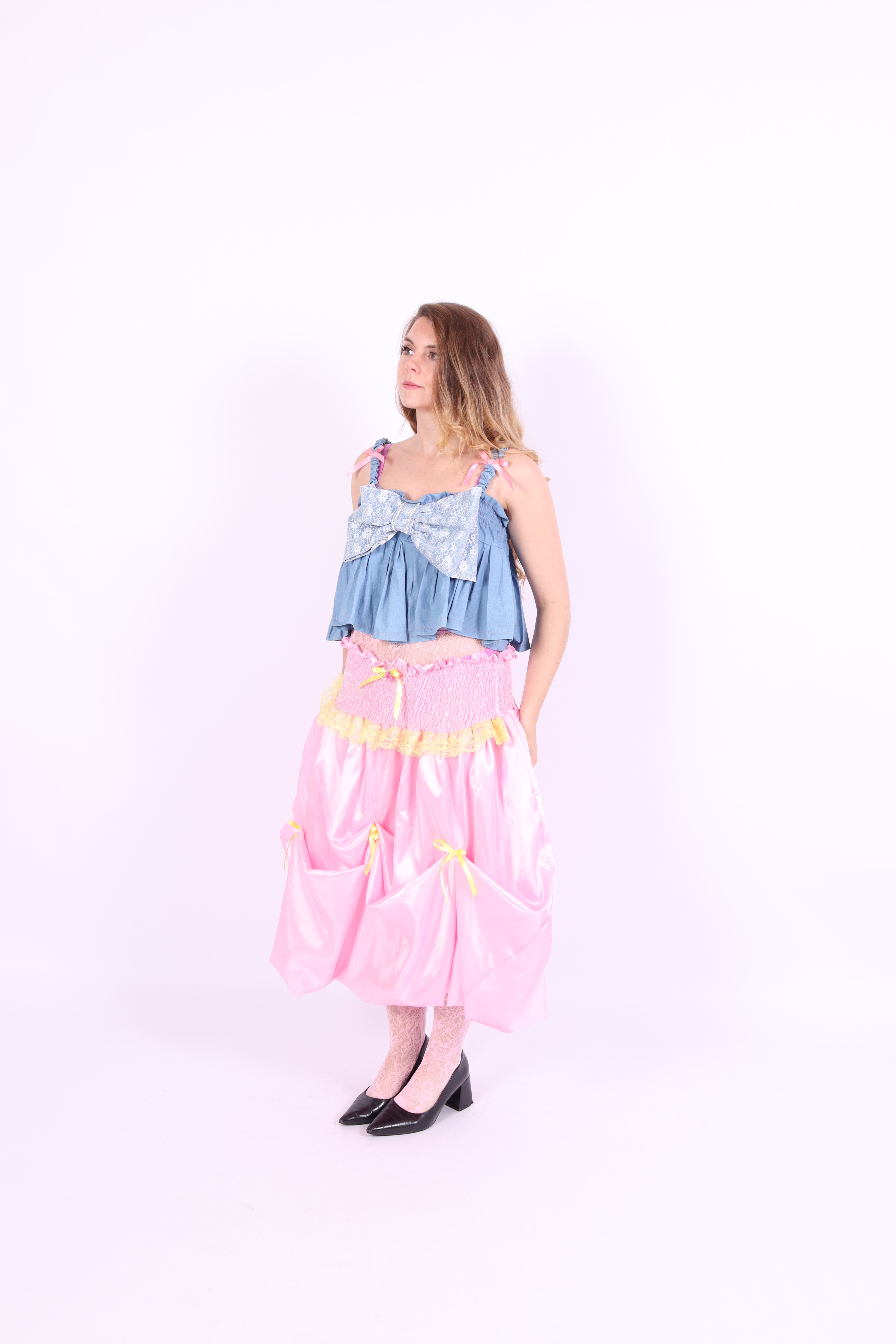 A denim crop top with a large white lace bow and diamante detail on the chest, pink topstitch elasticated shirring across the top bodice, and full babydoll gathered panel. It features elasticated shoulder straps with pink ribbon bow detail. Delta Of Phoenix Gigi Bow Top