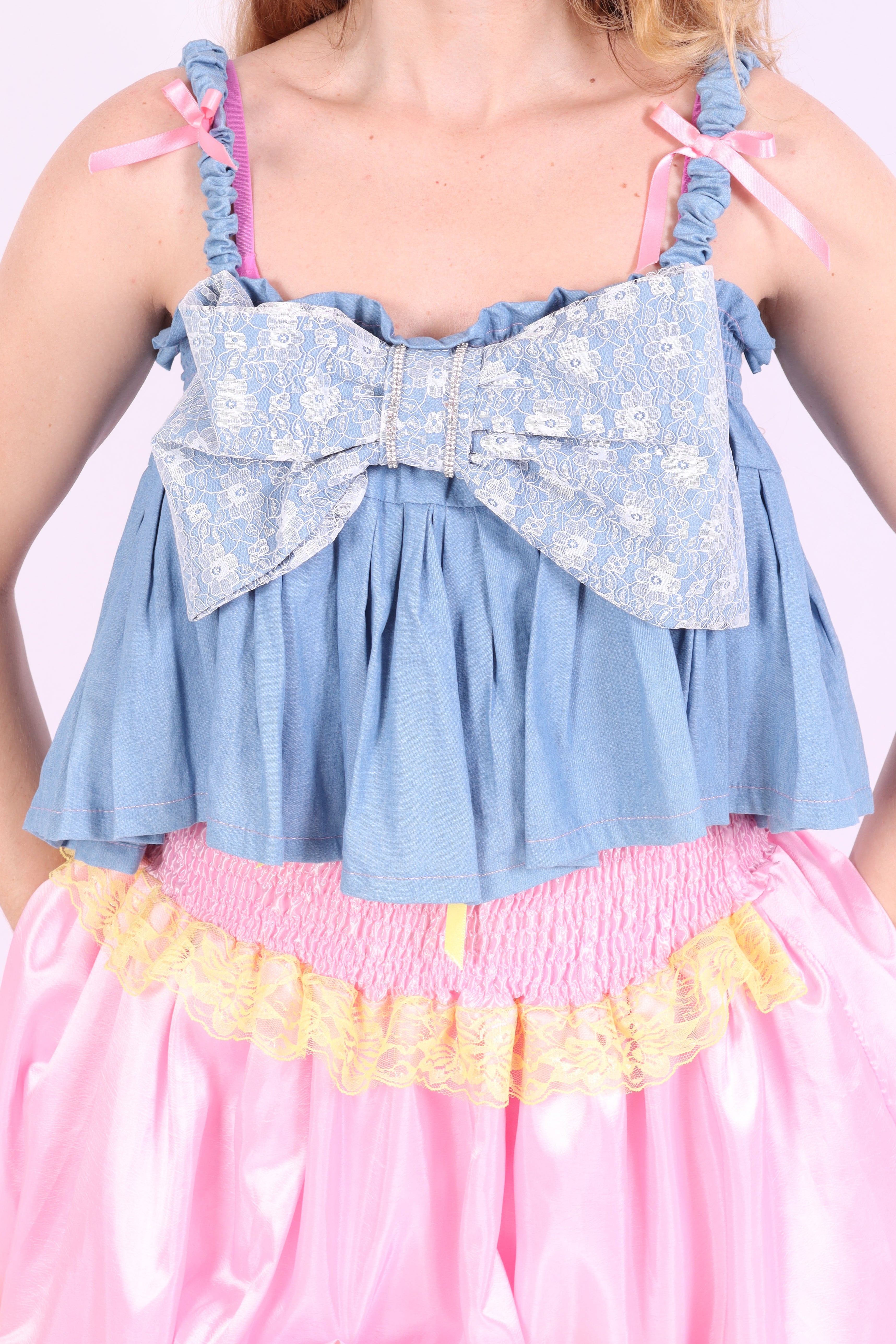 A denim crop top with a large white lace bow and diamante detail on the chest, pink topstitch elasticated shirring across the top bodice, and full babydoll gathered panel. It features elasticated shoulder straps with pink ribbon bow detail. Delta Of Phoenix Gigi Bow Top
