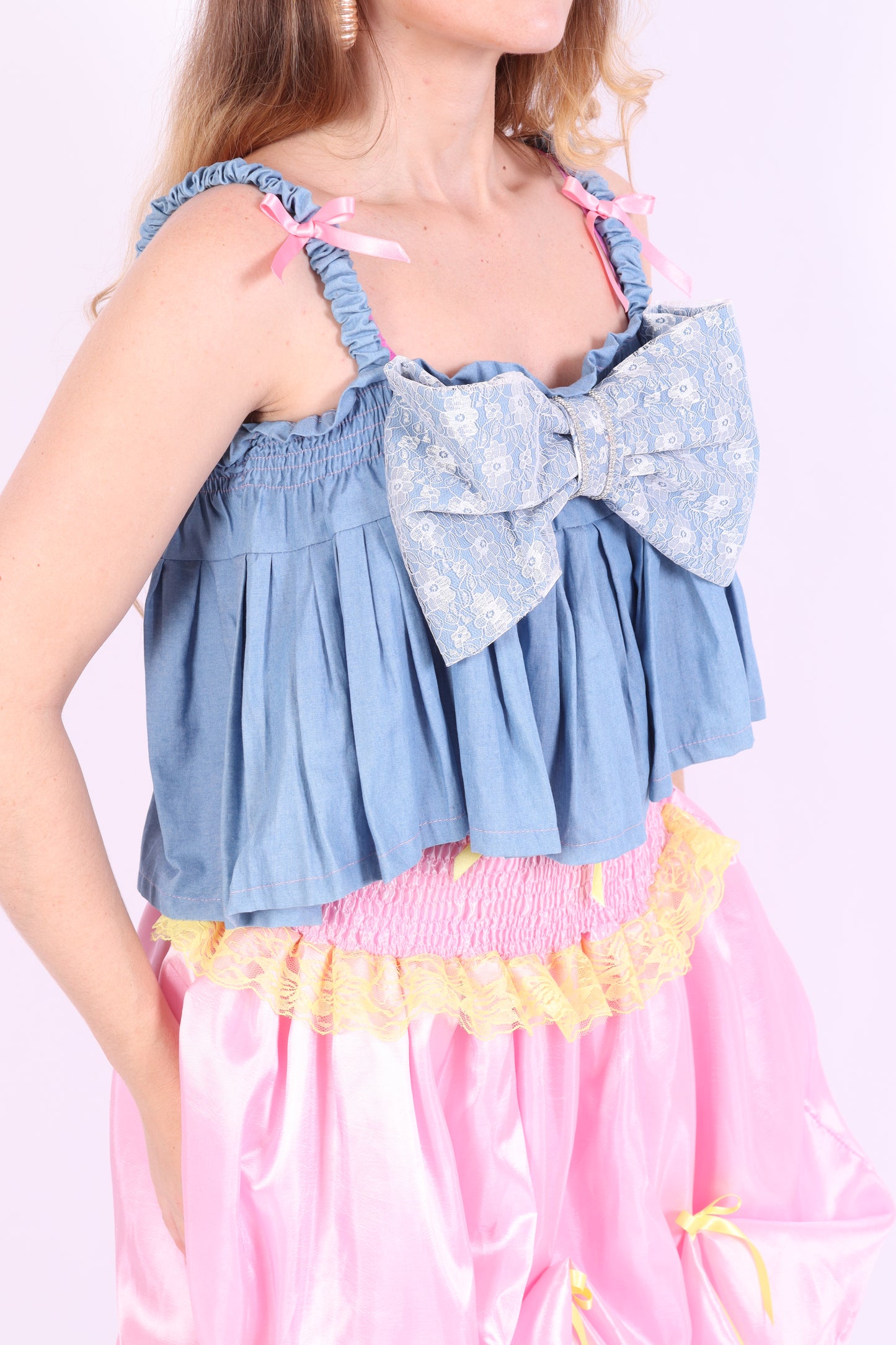 A denim crop top with a large white lace bow and diamante detail on the chest, pink topstitch elasticated shirring across the top bodice, and full babydoll gathered panel. It features elasticated shoulder straps with pink ribbon bow detail. Delta Of Phoenix Gigi Bow Top